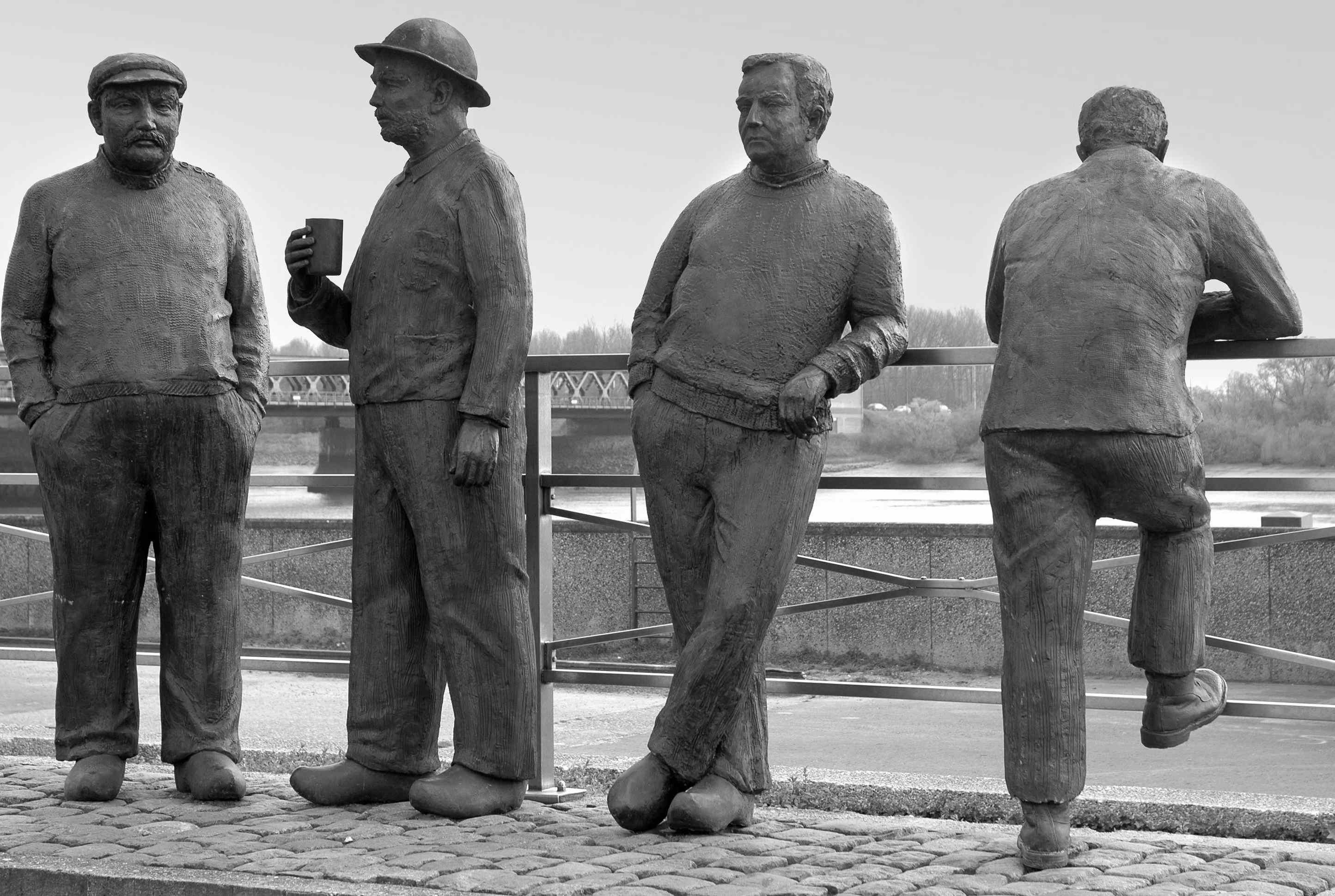 "Picture of statues of four working class men leaning on a fence"