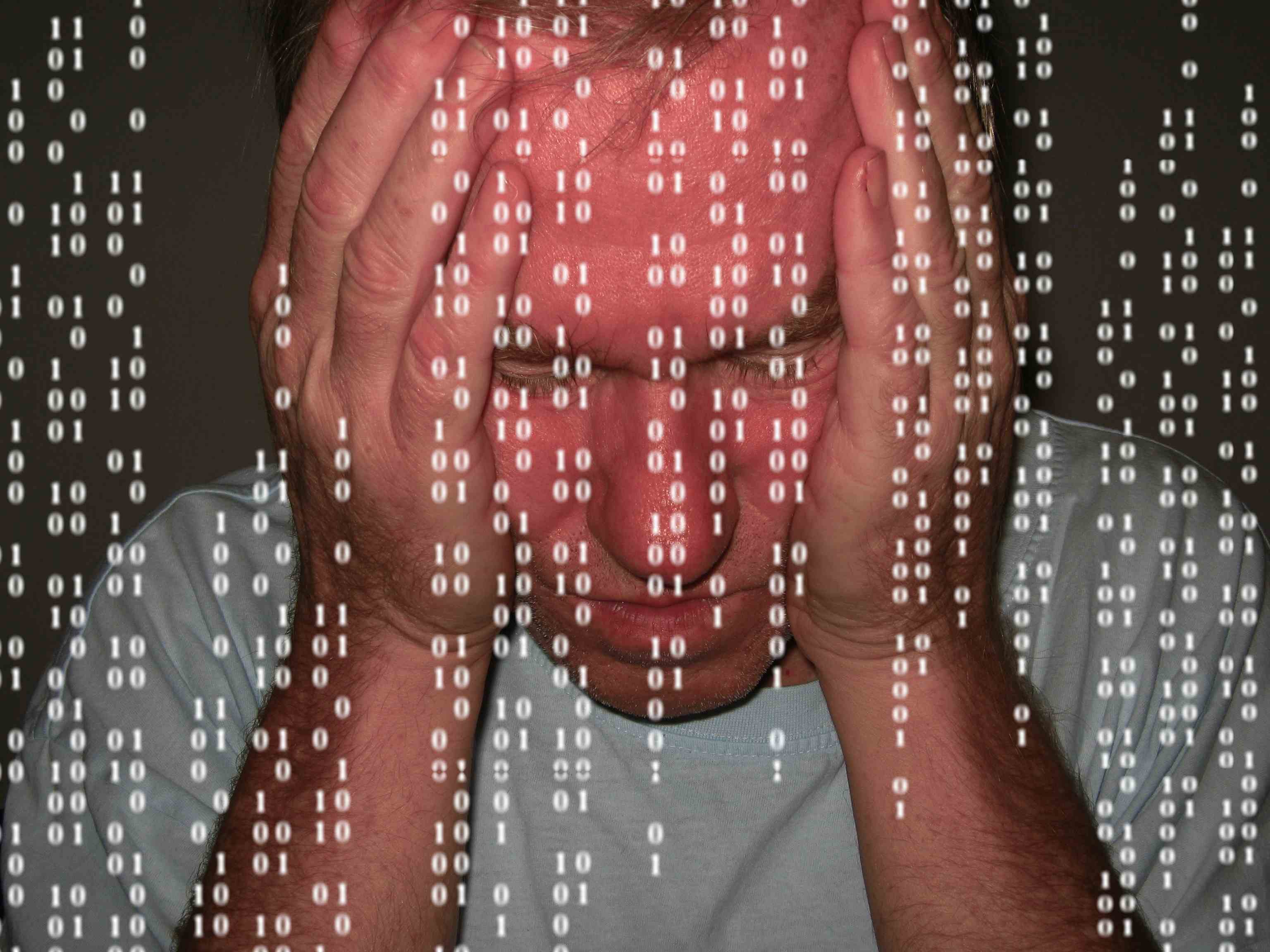 Man looking overwhelmed, holding head in hands, screen covered with binary digits