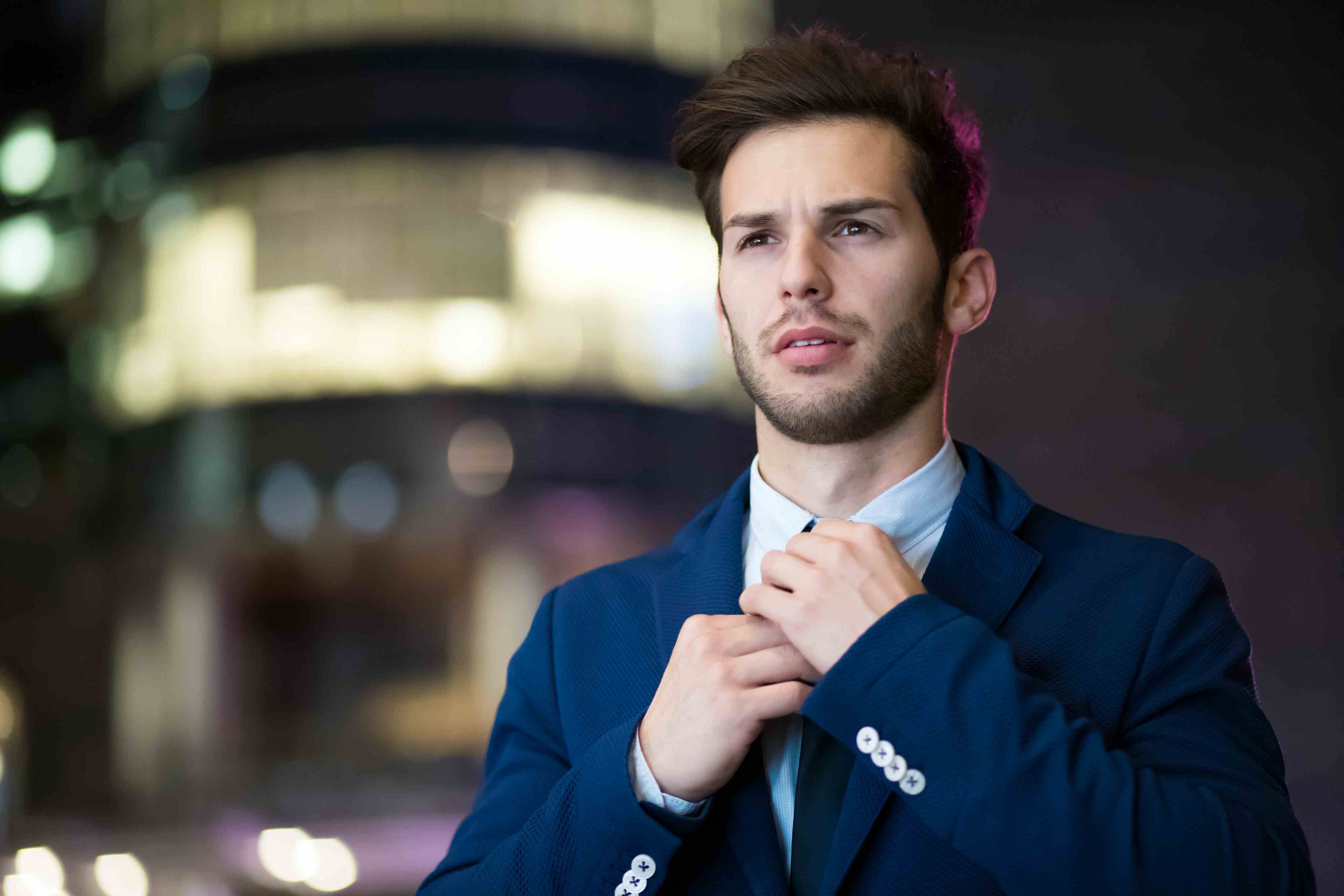 Young, handsome man, in tailored blue suit, straightening his tie with neon lights in background